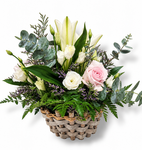 Arrangement floral pastel dans un panier tressé, composé de lys blancs, de roses rose pâle, de lisianthus crème et de feuillage d’eucalyptus.