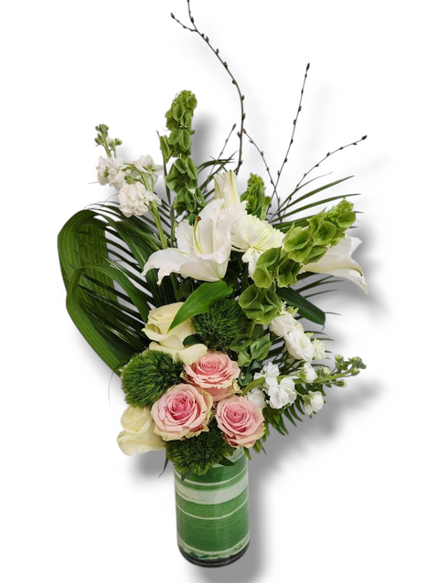 Arrangement floral vertical avec lys blancs, cloches d’Irlande, roses roses et feuillage tropical dans vase vert décoratif.
