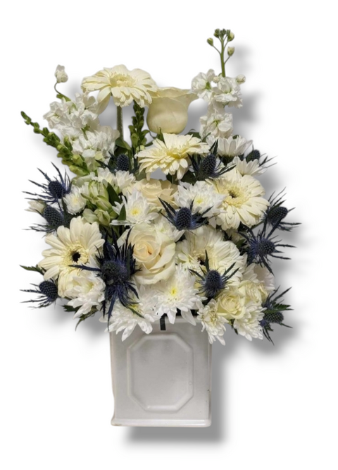 Arrangement funéraire blanc et bleu avec roses, gerberas, chardons et fleurs blanches dans un contenant blanc.