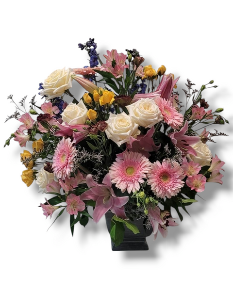 Arrangement floral pastel et coloré avec roses crème, germinis roses, lys, delphiniums et feuillage – modèle Jardin d’Émotions.