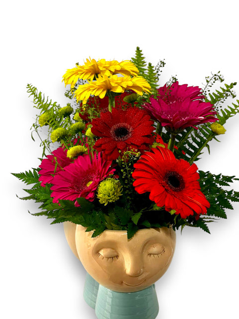 Arrangement floral avec germinis rouges, jaunes et fuchsia dans un vase en céramique à visage souriant – modèle « Sourire en Pot » de Fleur de Guy.