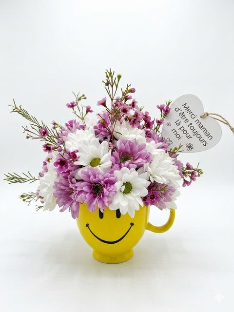 Arrangement floral fête des Mères avec marguerites blanches et mauves dans une tasse jaune sourire, message cœur pour maman.