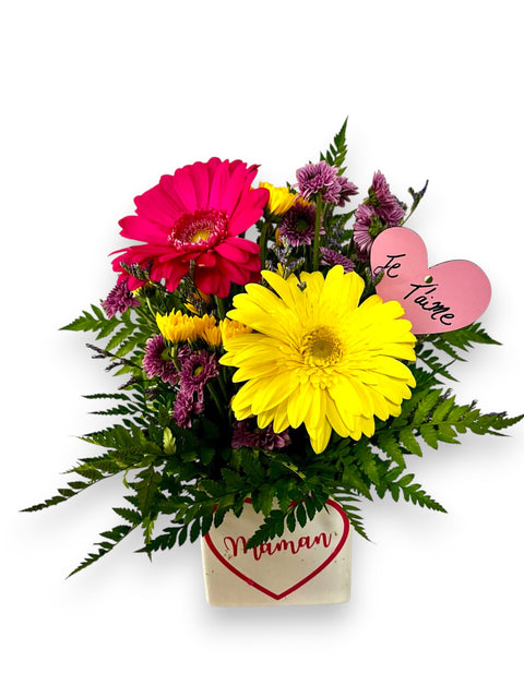 Arrangement floral Fête des Mères avec germinis jaunes et roses dans un pot avec cœur « Maman » et cœur rose « Je t’aime ».