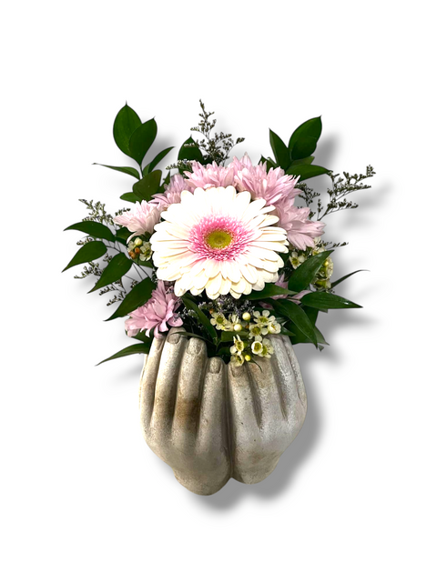 Arrangement floral pastel dans vase en forme de mains – gerbera rose pâle, petites fleurs blanches et feuillage tendre.
