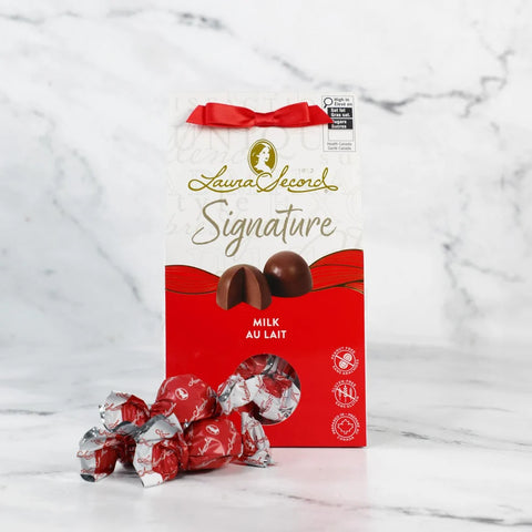 Boîte de chocolats Laura Secord Signature au lait avec ruban rouge et chocolats emballés individuellement, présentée sur un fond de marbre blanc.