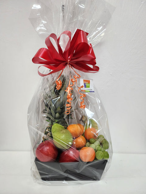 Panier cadeau de fruits variés avec ananas, pommes, poire, raisins, oranges, décoré d’une boucle rouge et emballé dans du cellophane.