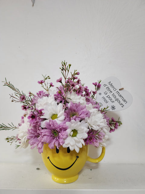 Arrangement floral fête des Mères avec marguerites blanches et mauves dans une tasse jaune sourire, message cœur pour maman.