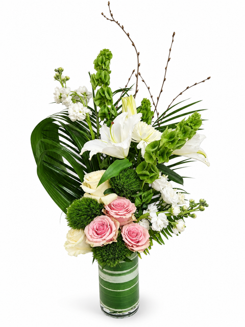 Arrangement floral blanc, vert et rose pâle composé de lys blancs, cloches d’Irlande, roses, feuillage tropical et branches décoratives dans un vase cylindrique vert.
