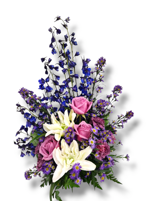 Arrangement floral apaisant avec lys blancs, roses mauves et delphinium bleu en forme pyramidale.

