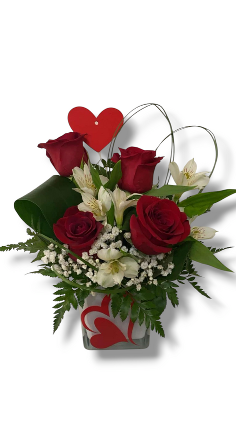 Arrangement floral de la Saint-Valentin avec roses rouges, fleurs blanches et cœurs décoratifs dans un vase carré.