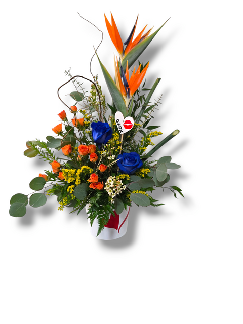 Arrangement floral exotique avec oiseaux du paradis, roses bleues, mini-roses orange et feuillage – dans un vase décoré d’un cœur rouge.