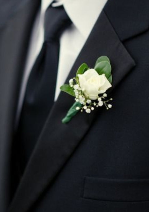 Boutonnière chic avec rose blanche, feuillage vert et gypsophile sur complet noir.

