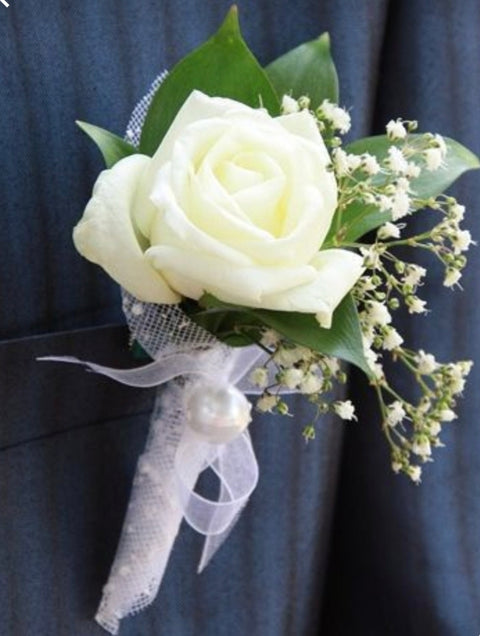 Boutonnière avec rose blanche, gypsophile et ruban blanc perlé, portée sur veston foncé.

