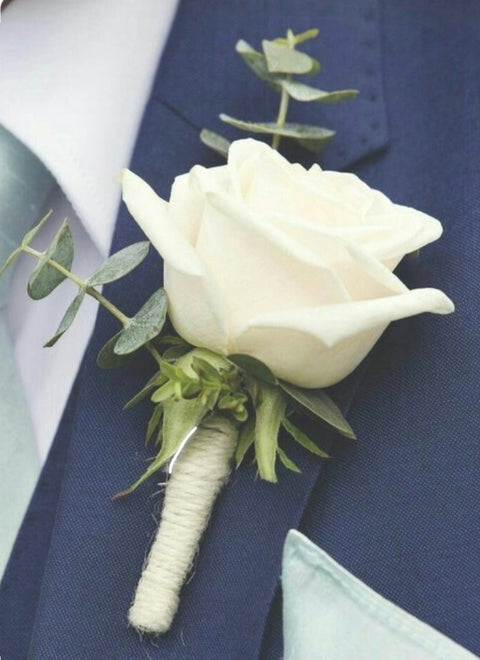 Boutonnière avec rose ivoire, eucalyptus et tige enroulée de corde, portée sur complet bleu.

