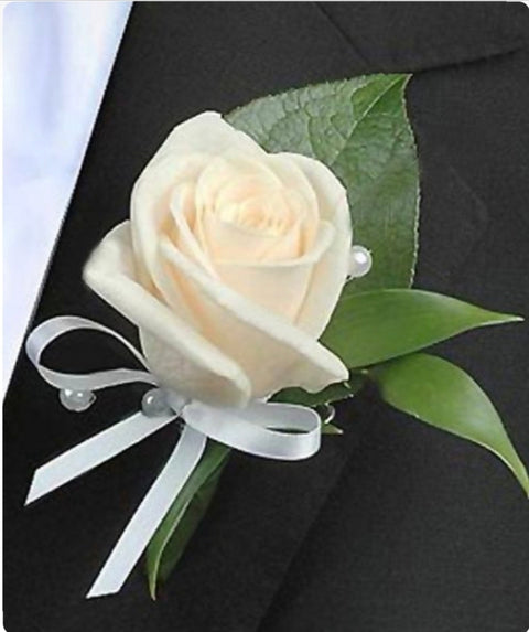 Boutonnière avec rose ivoire, feuillage vert, ruban blanc satiné et perles, portée sur un complet noir.

