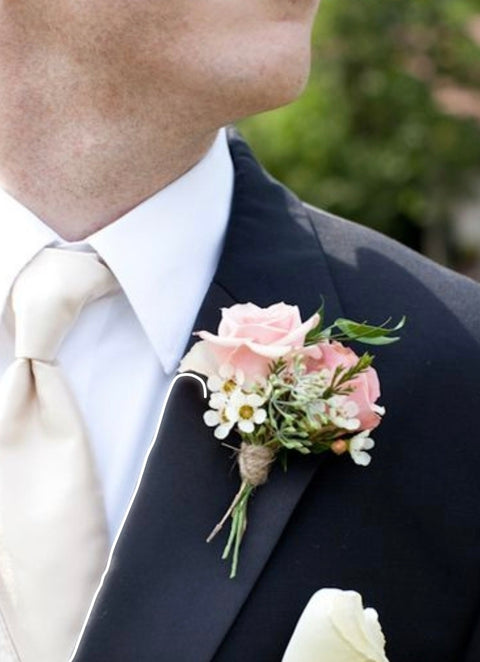 Boutonnière avec mini-roses rose pâle, marguerites blanches et ficelle de jute, portée par un homme en complet.

