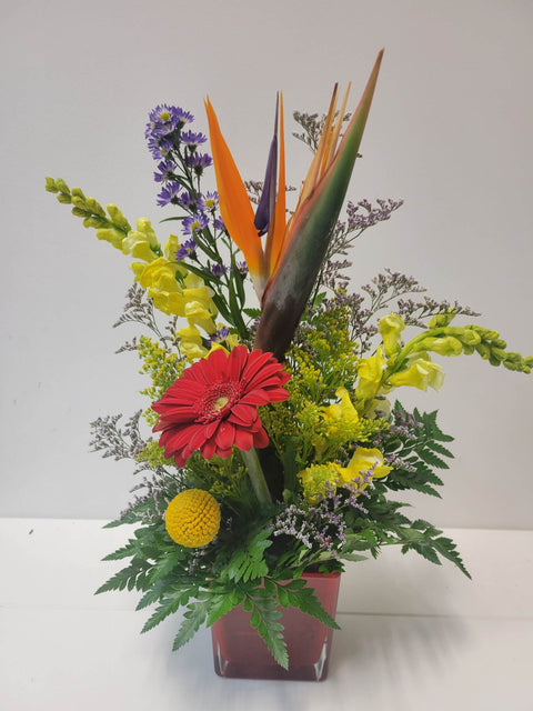 Arrangement floral tropical avec oiseau du paradis, gerbera rouge, fleurs jaunes et violettes dans un vase rouge.