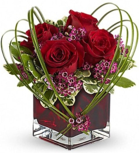 Arrangement floral romantique avec roses rouges et feuillage en forme de cœur dans un vase cube.

